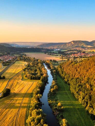 Die Saale aus der Vogelperspektive im Licht der Abendsonne. Links des Flusses liegen Felder, rechts ein Wald. Am Horizont liegt die Leuchtenburg.