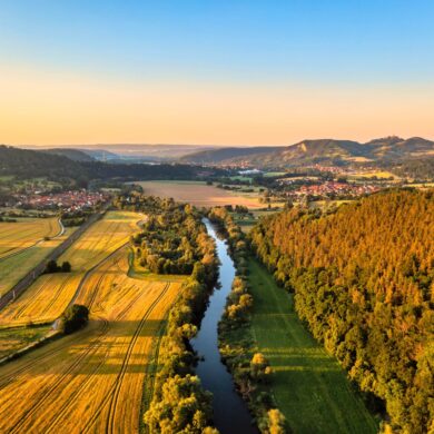 Die Saale aus der Vogelperspektive im Licht der Abendsonne. Links des Flusses liegen Felder, rechts ein Wald. Am Horizont liegt die Leuchtenburg.