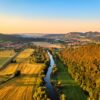 A bird's eye view of the Saale in the light of the evening sun. To the left of the river are fields, to the right a forest. Leuchtenburg Castle lies on the horizon.