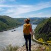 Eine Frau mit Wanderrucksack steht auf einem Felsvorsprung, unter ihr eine Rheinschleife mit Frachtschiff