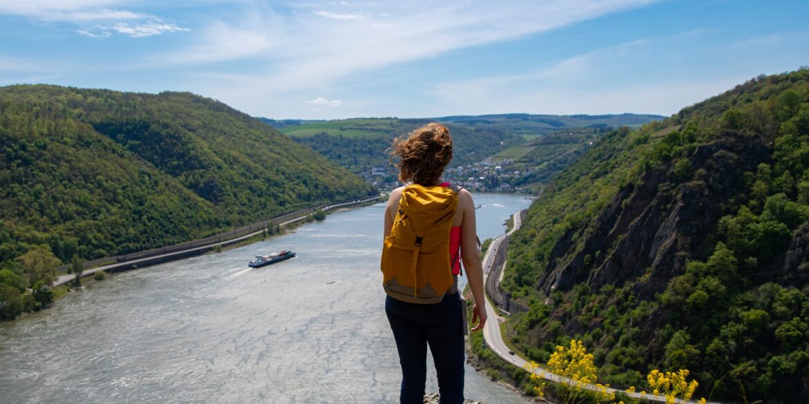 Eine Frau mit Wanderrucksack steht auf einem Felsvorsprung, unter ihr eine Rheinschleife mit Frachtschiff
