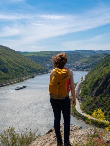 Eine Frau mit Wanderrucksack steht auf einem Felsvorsprung, unter ihr eine Rheinschleife mit Frachtschiff