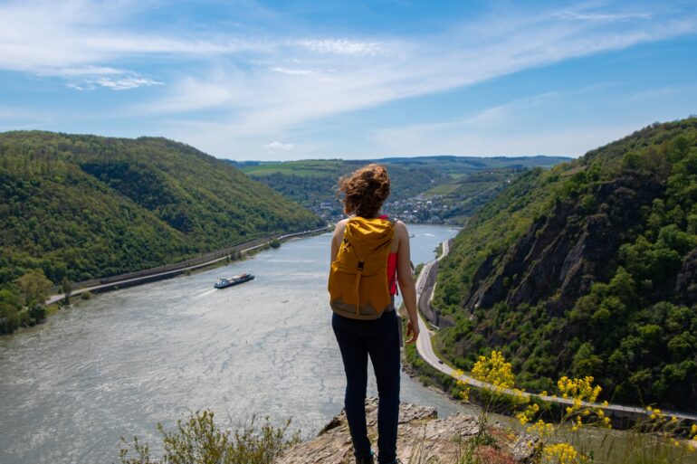 Eine Frau mit Wanderrucksack steht auf einem Felsvorsprung, unter ihr eine Rheinschleife mit Frachtschiff