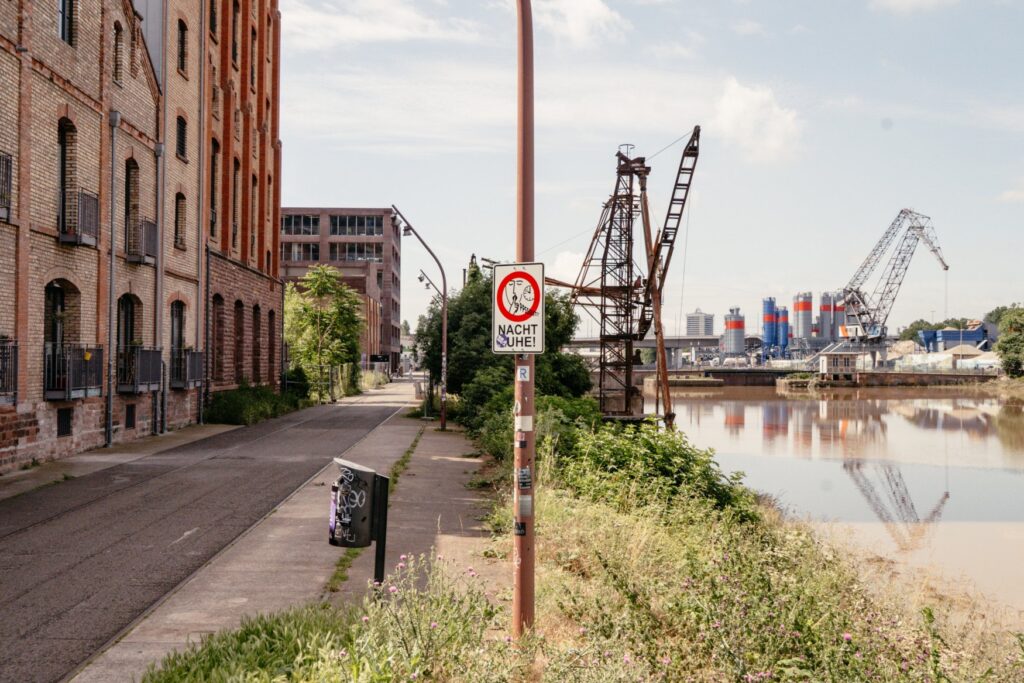 A sign reading “Please be quiet at night” stands on the shore of Mannheim harbor, with a brick building on the left