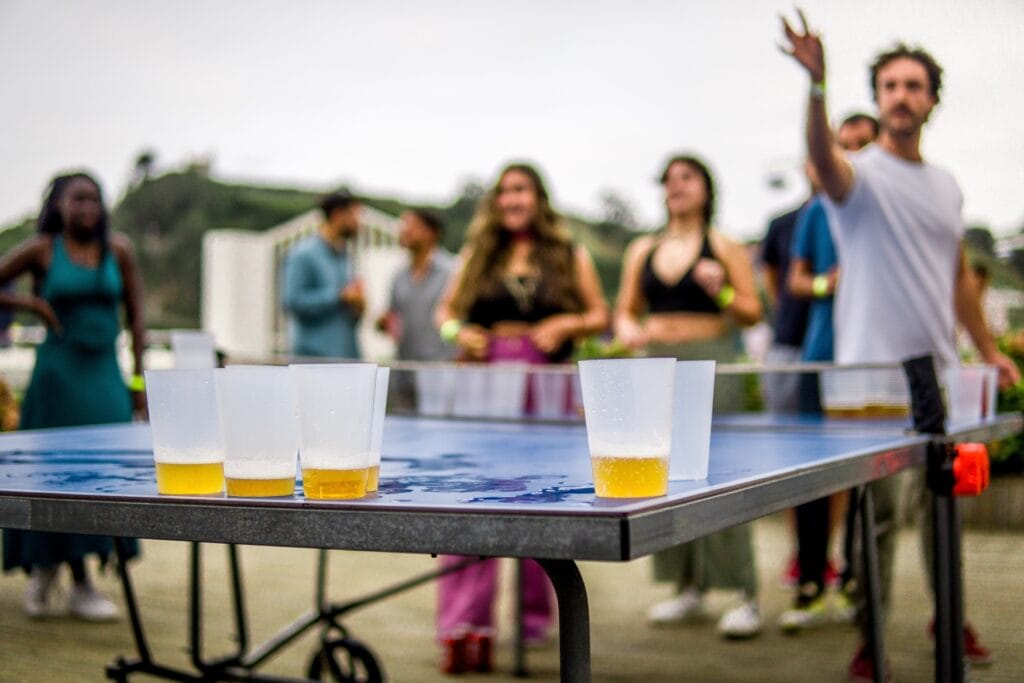 Auf einer Tischtennisplatte stehen einige halb mit Bier gefüllte Plastikbecher. Im Hintergrund ist unscharf ein Mann zu erkennen, der gerade zum Wurf ansetzt.