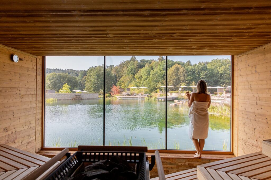 The sauna area at the wellness hotel Pfalzblick in the Palatinate Forest, with large panoramic windows overlooking the lake. A woman in a towel stands at the window and looks out.