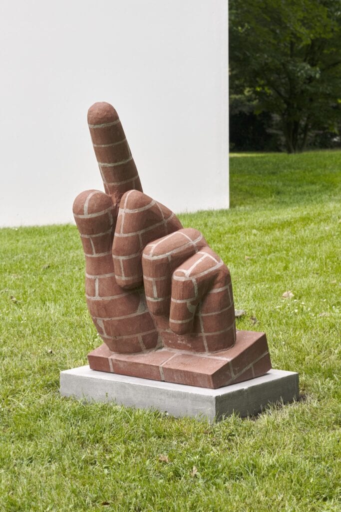 Eine steinerne Hand im Skulpturenpark Köln, die wie eine Ziegelwand bemalt ist. Der Zeigefinger ist in die Höhe gestreckt.