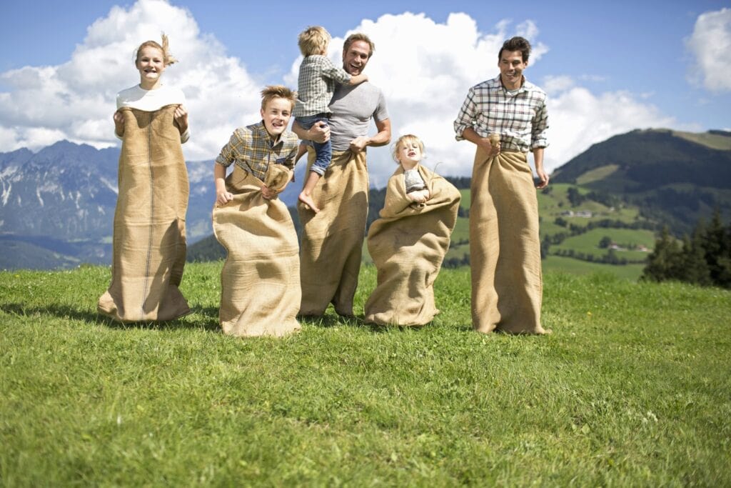 Zwei Männer und drei Kinder spielen Sackhüpfen auf einer sonnigen Alpenwiese, im Hintergrund Berge