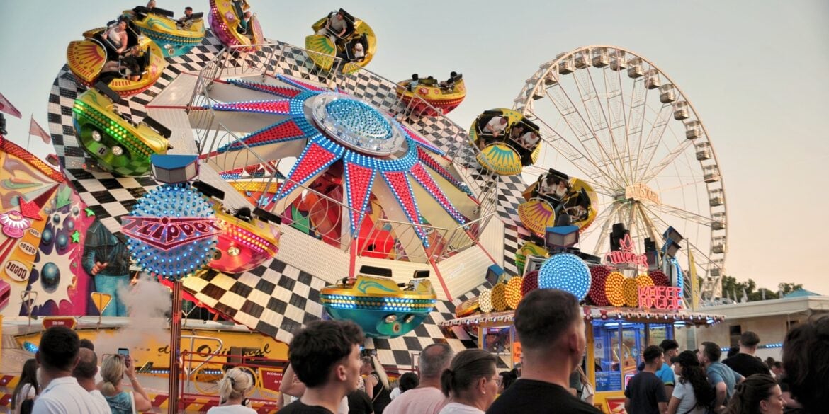 Ein buntes Fahrgeschäft auf einem deutschen Volksfest; mehrere Kabinen auf einem sich drehenden Rad, davor gehen einige Menschen vorbei. Im Hintergrund ist ein Riesenrad zu sehen.