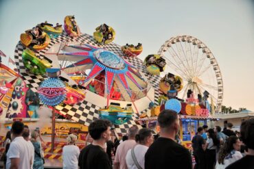 Ein buntes Fahrgeschäft auf einem deutschen Volksfest; mehrere Kabinen auf einem sich drehenden Rad, davor gehen einige Menschen vorbei. Im Hintergrund ist ein Riesenrad zu sehen.
