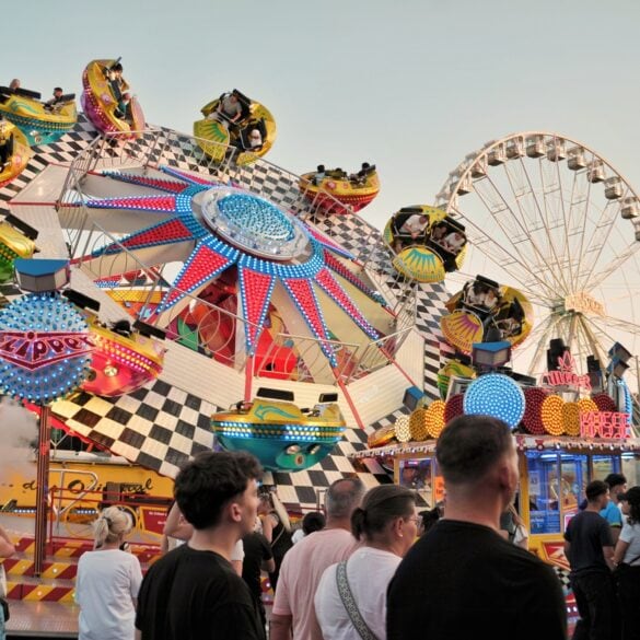 Ein buntes Fahrgeschäft auf einem deutschen Volksfest; mehrere Kabinen auf einem sich drehenden Rad, davor gehen einige Menschen vorbei. Im Hintergrund ist ein Riesenrad zu sehen.