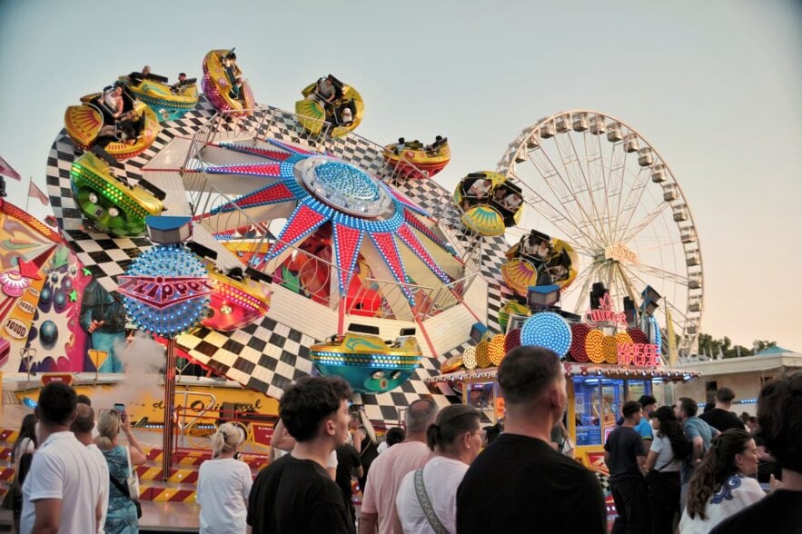 Ein buntes Fahrgeschäft auf einem deutschen Volksfest; mehrere Kabinen auf einem sich drehenden Rad, davor gehen einige Menschen vorbei. Im Hintergrund ist ein Riesenrad zu sehen.