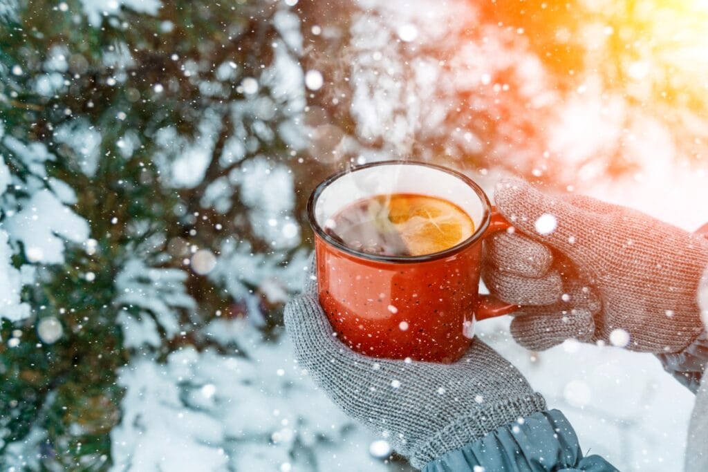 Eine Person hält eine dampfende Tasse Punsch in der Hand, dahinter sind verschneite Nadelbäume