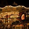 Ein reich verziertes Kinderkarussell auf einem Weihnachtsmarkt. Auf der obersten Ebene sind verschiedene Stadtansichten zu sehen, darunter das eigentliche Karussell, das mit vielen Tannenzweigen und Lichterketten ausgestattet ist. Im Vordergrund verdeckt eine kleine Hütte den Blick, das Bild wurde spät abends auf einem Weihnachtsmarkt aufgenommen.