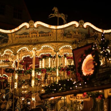 Ein reich verziertes Kinderkarussell auf einem Weihnachtsmarkt. Auf der obersten Ebene sind verschiedene Stadtansichten zu sehen, darunter das eigentliche Karussell, das mit vielen Tannenzweigen und Lichterketten ausgestattet ist. Im Vordergrund verdeckt eine kleine Hütte den Blick, das Bild wurde spät abends auf einem Weihnachtsmarkt aufgenommen.