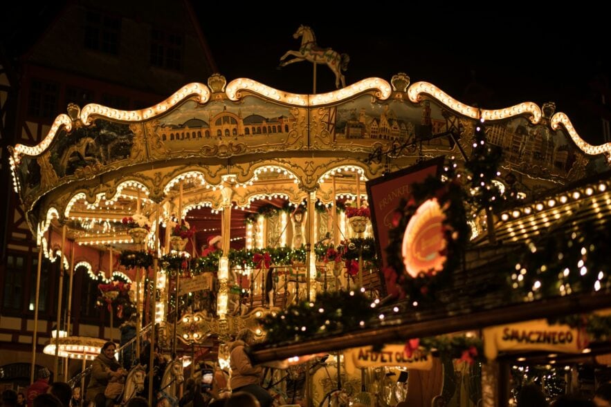 Ein reich verziertes Kinderkarussell auf einem Weihnachtsmarkt. Auf der obersten Ebene sind verschiedene Stadtansichten zu sehen, darunter das eigentliche Karussell, das mit vielen Tannenzweigen und Lichterketten ausgestattet ist. Im Vordergrund verdeckt eine kleine Hütte den Blick, das Bild wurde spät abends auf einem Weihnachtsmarkt aufgenommen.
