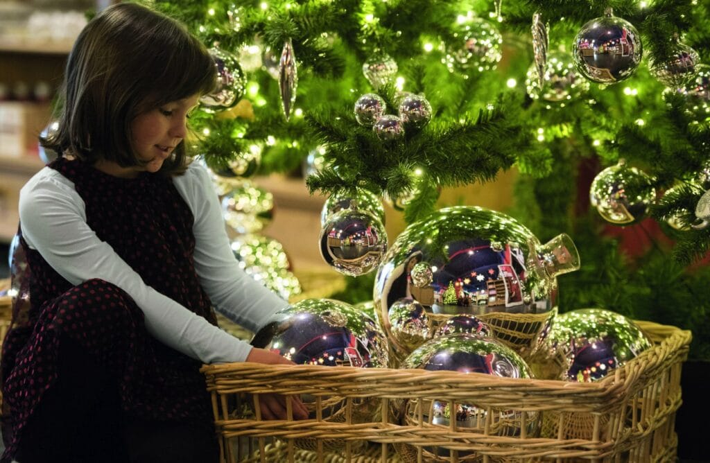 Ein Kind bewundert mehrere sehr große Weihnachtsbaumkugeln in einem Korb unter einem geschmückten Baum