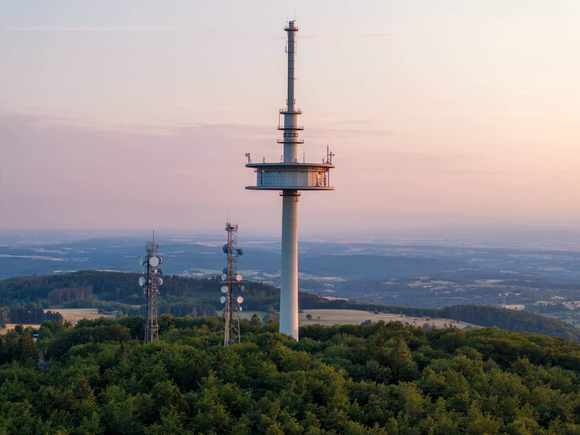 Drohnenaufnahme des Funkturms auf dem Vogelsberg in Hessen, im Hintergrund scheint sanft das Licht der Abenddämmerung