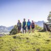 Vier Menschen wandern über eine grüne Hochwiese, im Hintergrund ein Alpenkamm