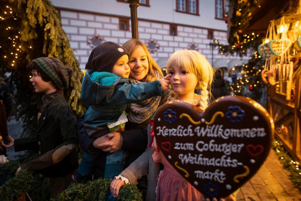 Toddler in her mother's arms admires a doll holding a gingerbread heart with the inscription “Welcome to the Coburg Christmas market”