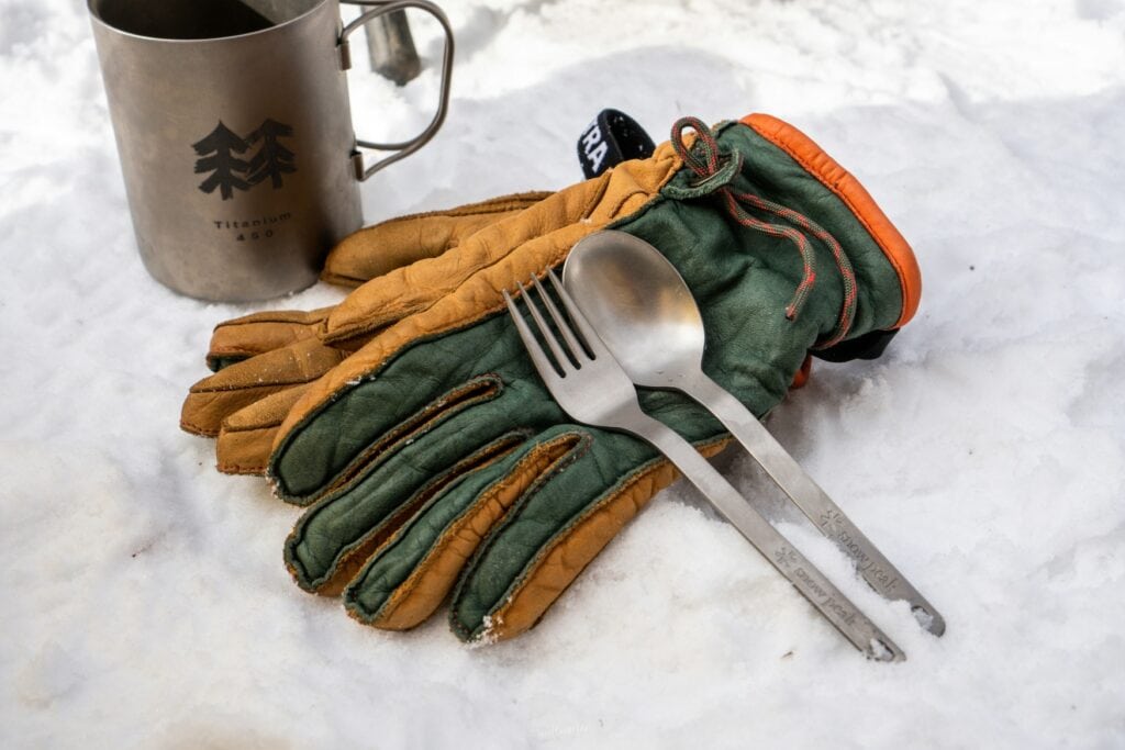 Eine Metalltasse, Besteck und ein paar Handschuhe liegen auf Schnee