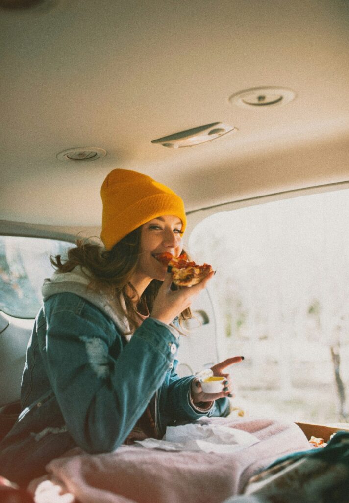 A woman in winter clothes eats a slice of pizza in a camper van. Outside, a snowy landscape