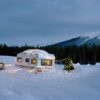 A snow-covered caravan is wrapped in a string of lights, next to it stands a Christmas tree.