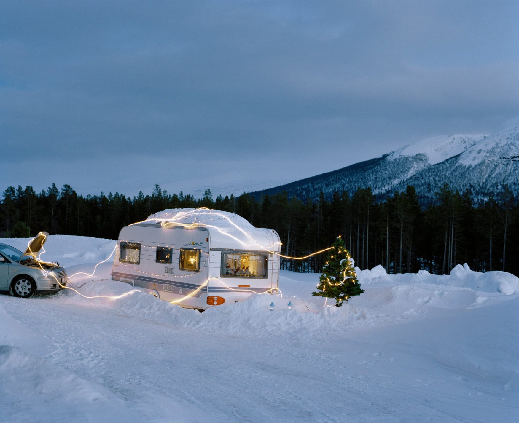 A snow-covered caravan is wrapped in a string of lights, next to it stands a Christmas tree.