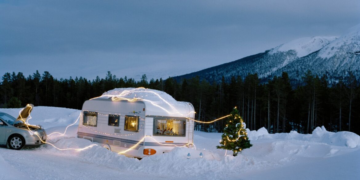 Ein überschneiter Wohnwagen ist mit einer Lichterkette umwickelt, daneben steht ein Weihnachtsbaum