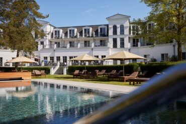 Außenbereich des Strandhotels Zingst im Sonnenschein, im Vordergrund ein Pool und einige Liegen mit Sonnenschirm