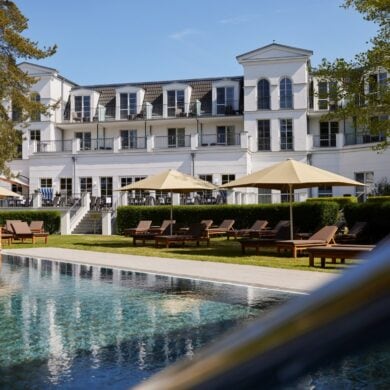 Außenbereich des Strandhotels Zingst im Sonnenschein, im Vordergrund ein Pool und einige Liegen mit Sonnenschirm