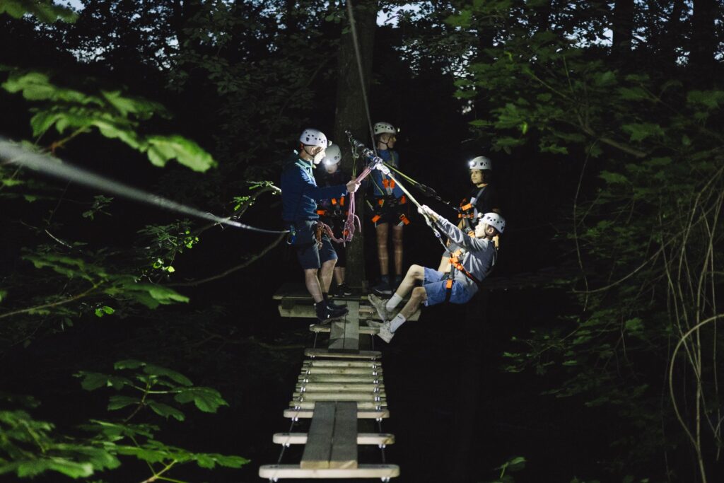 Eine Gruppe Menschen balanciert über eine Brücke in einem nächtlichen Kletterwald