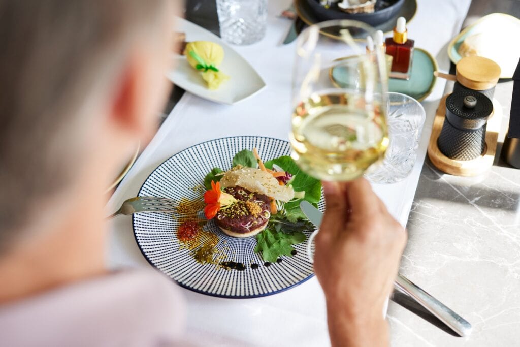 Ein elegant angerichteter Teller mit einem Tartar-Burger. Im Vordergrund ist unscharf ein Mann zu erkennen, der ein Glas Weißwein in der Hand hält