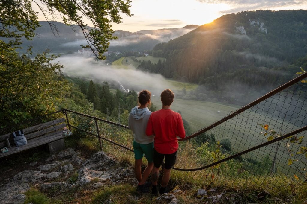 Zwei Personen schauen sich den Sonnenaufgang über einem nebeligen Tal im Donaubergland an