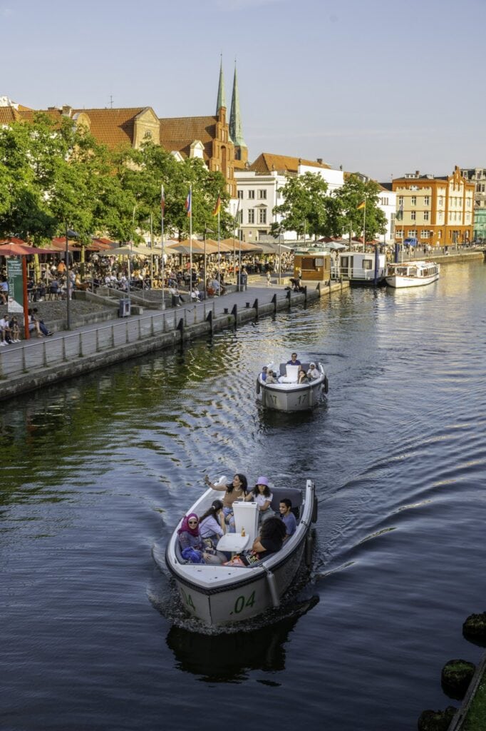 Kleine Ausflugsboote auf der Trave in Lübeck. Am Ufer sitzen Menschen in Außengastronomien