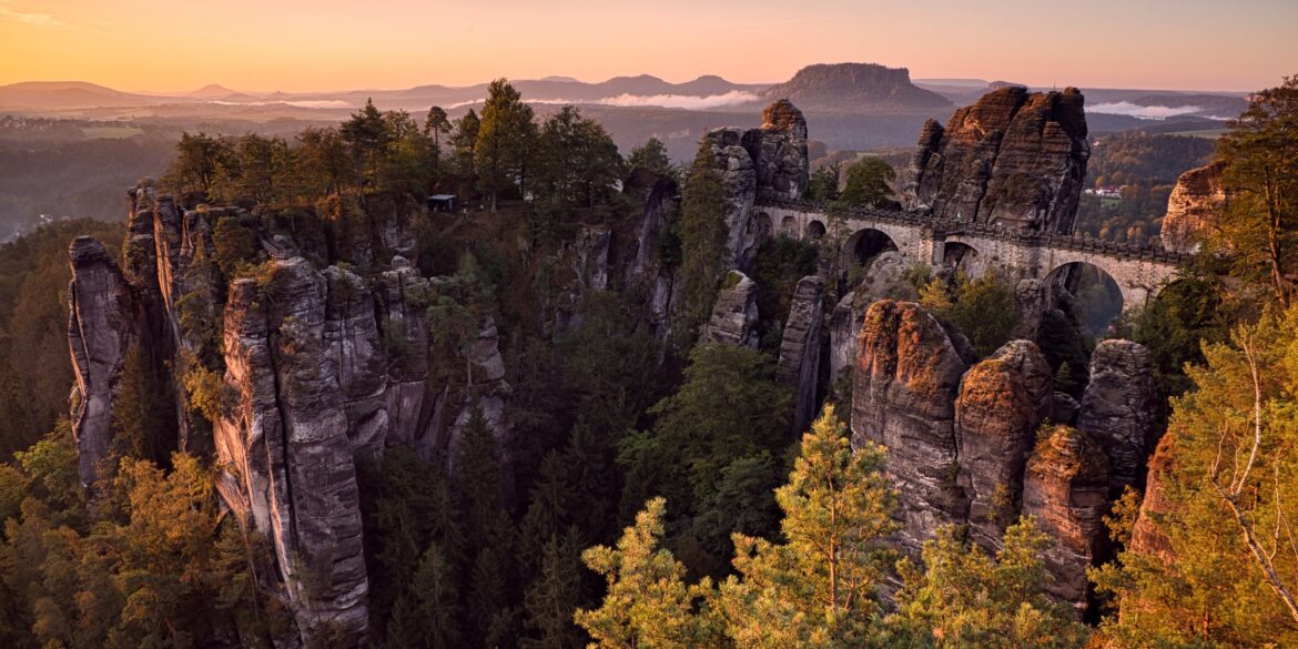 Über ein Felsenmeer spannt eine mächtige Sandsteinbrücke vom rechten Bildrand bis zu einem mittigen Felsplateau. Dahinter eine weite Ebene, über der gerade die Herbstsonne an einem klaren Himmel untergeht.