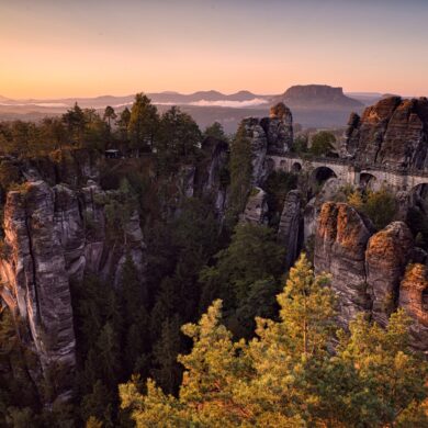 Über ein Felsenmeer spannt eine mächtige Sandsteinbrücke vom rechten Bildrand bis zu einem mittigen Felsplateau. Dahinter eine weite Ebene, über der gerade die Herbstsonne an einem klaren Himmel untergeht.