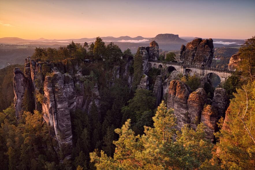 Über ein Felsenmeer spannt eine mächtige Sandsteinbrücke vom rechten Bildrand bis zu einem mittigen Felsplateau. Dahinter eine weite Ebene, über der gerade die Herbstsonne an einem klaren Himmel untergeht.