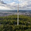 Drohnenaufnahme des Stuttgarter Fernsehturms, der aus einem grünen Waldstück in die Höhe ragt. Hinter dem Wald erstreckt sich die Stadt über das ganze Bild, darüber ein teilweise bewölkter Himmel.
