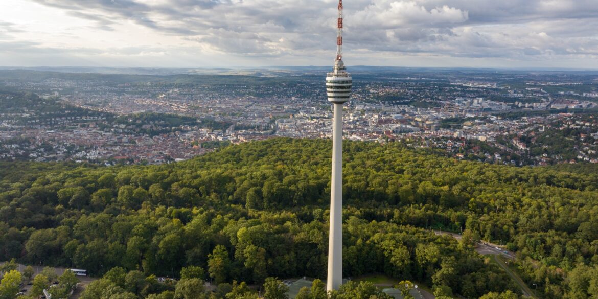 Drohnenaufnahme des Stuttgarter Fernsehturms, der aus einem grünen Waldstück in die Höhe ragt. Hinter dem Wald erstreckt sich die Stadt über das ganze Bild, darüber ein teilweise bewölkter Himmel.