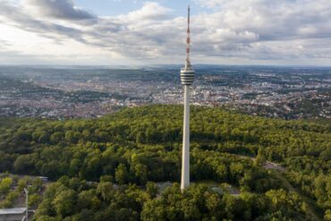 Drohnenaufnahme des Stuttgarter Fernsehturms, der aus einem grünen Waldstück in die Höhe ragt. Hinter dem Wald erstreckt sich die Stadt über das ganze Bild, darüber ein teilweise bewölkter Himmel.