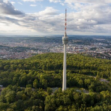 Drohnenaufnahme des Stuttgarter Fernsehturms, der aus einem grünen Waldstück in die Höhe ragt. Hinter dem Wald erstreckt sich die Stadt über das ganze Bild, darüber ein teilweise bewölkter Himmel.