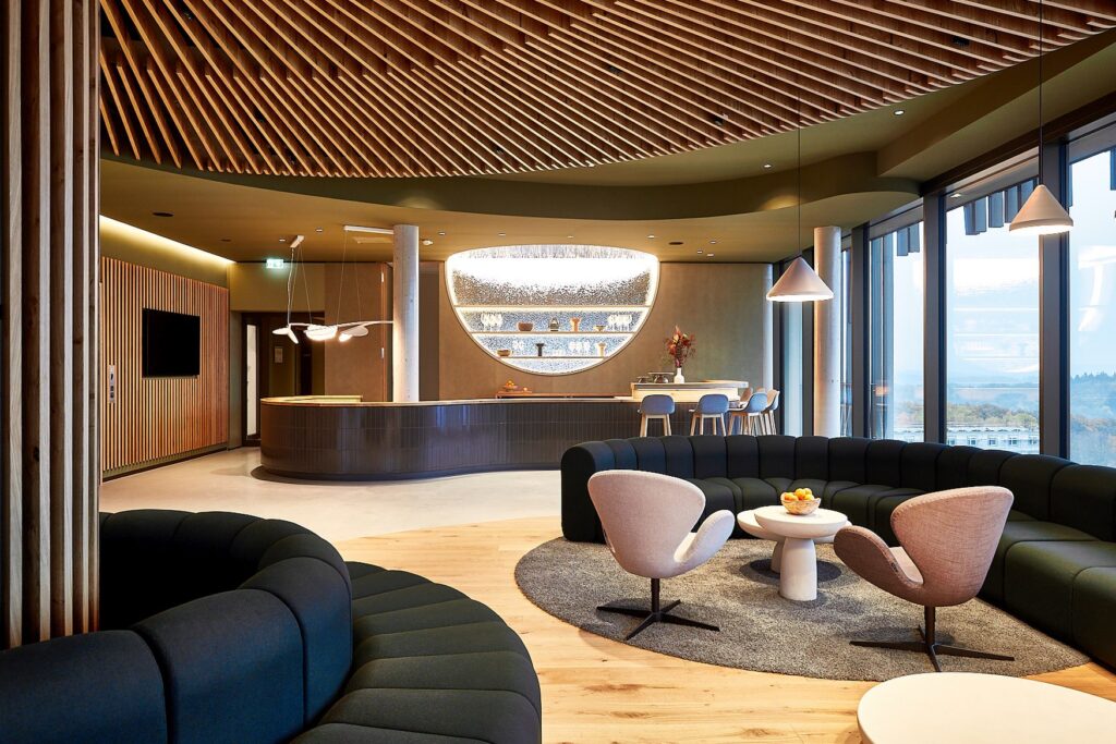 Lobby of the resort-hotel Wald.Weit, with softly curved upholstered furniture, wooden lamellas on the ceiling and a wide panoramic view