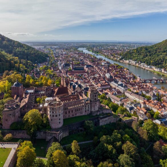 Drohnenansicht der Stadt Heidelberg, links im Vordergrund das Schloss, darunter im Tal die Altstadt und dann der Neckar. Aufgenommen an einem sonnigen Sommertag.