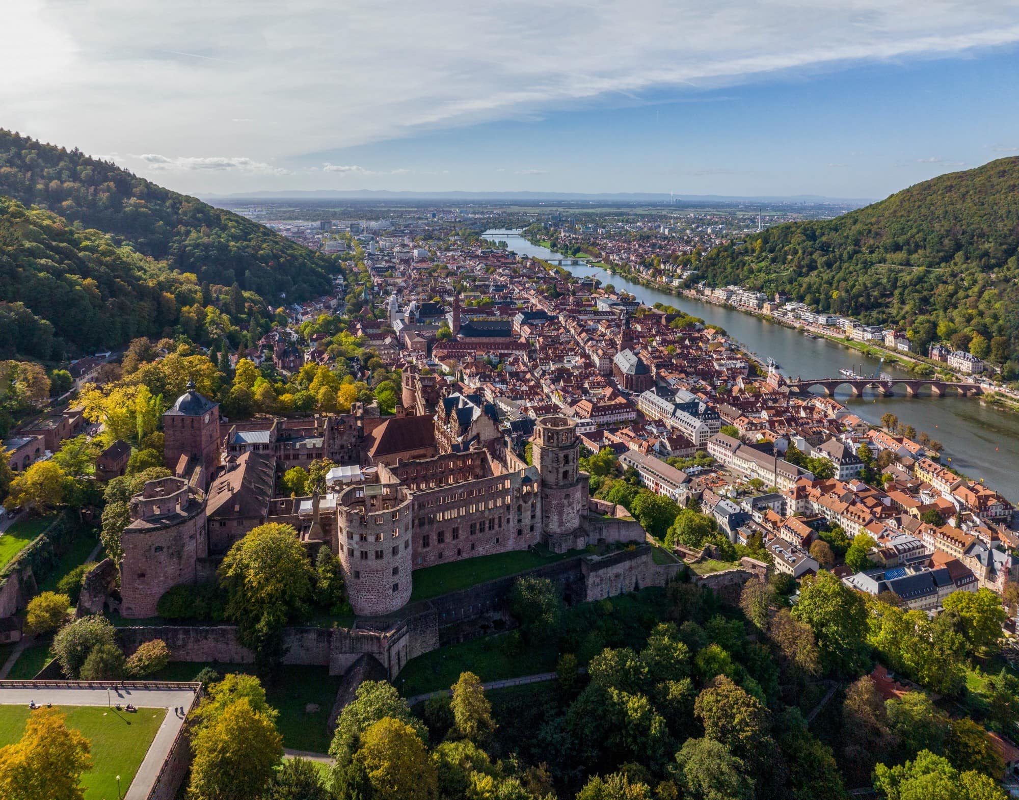 Drohnenansicht der Stadt Heidelberg, links im Vordergrund das Schloss, darunter im Tal die Altstadt und dann der Neckar. Aufgenommen an einem sonnigen Sommertag.