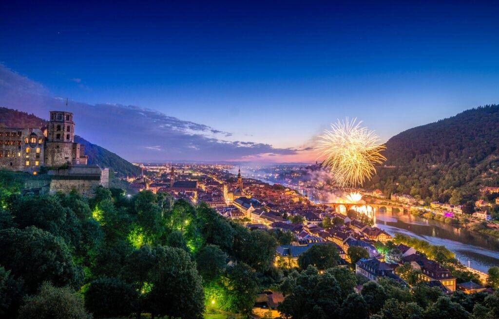 Drohnenansicht der Stadt Heidelberg, links im Vordergrund das Schloss, darunter im Tal die Altstadt und dann der Neckar im Abendlicht. Über dem Fluss explodiert gerade ein Feuerwerk.