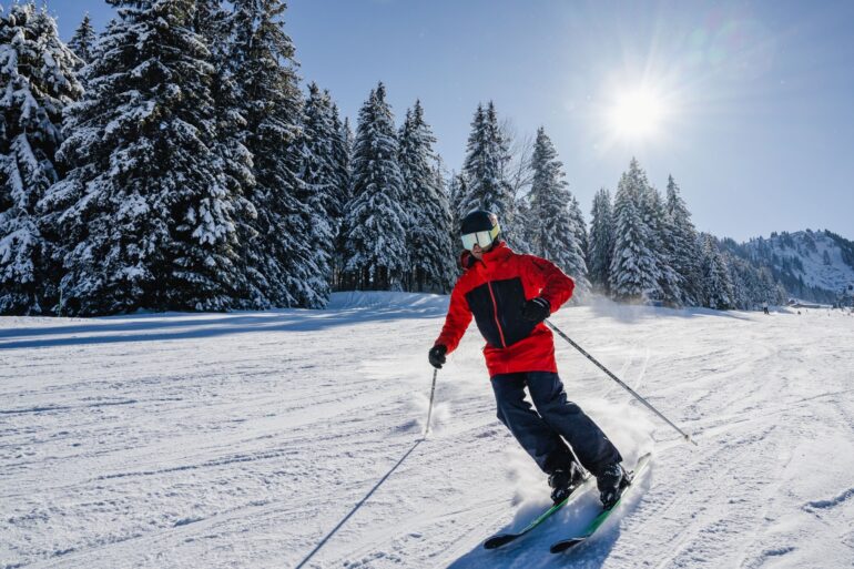 Ein Mann in roter Winterkleidung fährt auf Skiern eine Pista entlang. Die Bäume hinter der Piste sind dick mit Schnee verhangen, vom Himmel strahlt die Sonne hinab.