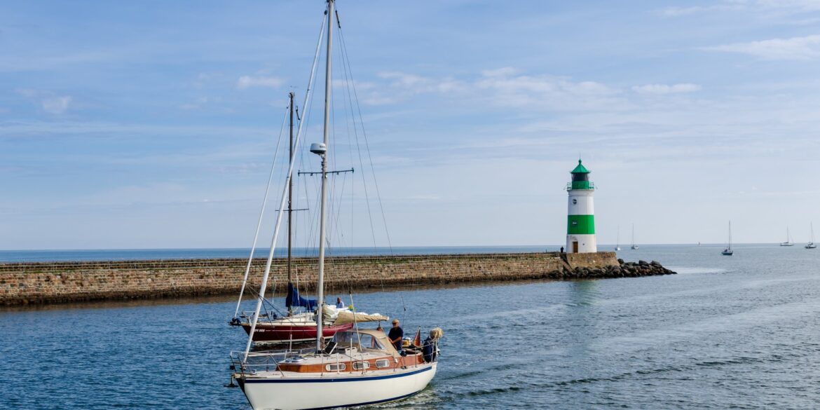 Zwei Segelboote fahren vor dem grün-weißen Leuchtturm in Kappeln an der Ostseeküste übers Wasser.