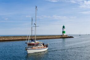 Zwei Segelboote fahren vor dem grün-weißen Leuchtturm in Kappeln an der Ostseeküste übers Wasser.