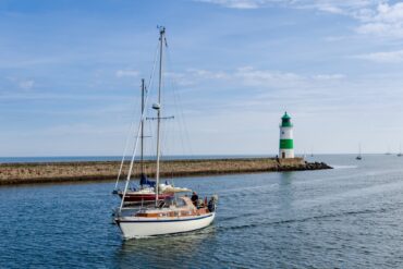 Zwei Segelboote fahren vor dem grün-weißen Leuchtturm in Kappeln an der Ostseeküste übers Wasser.